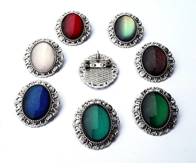 May include: Eight silver oval brooches with different colored glass stones. The stones are iridescent and change color in the light. The brooches are ornate and have a vintage style.