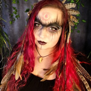 May include: A woman with long, curly red hair wears a headdress with feathers and a necklace with a bone pendant. She has dark eye makeup and a crescent moon painted on her forehead.
