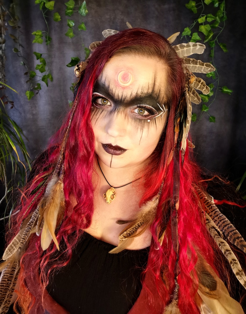 May include: A woman with long, curly red hair wears a headdress with feathers and a necklace with a bone pendant. She has dark eye makeup and a crescent moon painted on her forehead.