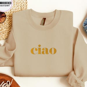 May include: A beige sweatshirt with the word "ciao" in gold lettering. The sweatshirt has a crew neck and long sleeves. The image also includes a pair of sunglasses, a pair of brown shoes, and a denim garment.