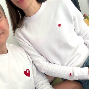 Op de afbeelding: Witte sweatshirts met rode geborduurde details. Eén heeft een rode puzzelstuk, de andere een rood hart met een puzzelstuk. De mouw van één sweatshirt heeft een rood hart en de letter "M".