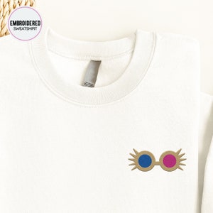 May include: White sweatshirt with embroidered gold-colored glasses with blue and pink lenses. The glasses have a whimsical design with eyelashes.