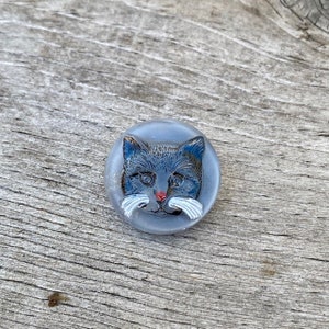 May include: A blue glass button with a painted image of a cat's face. The cat has white whiskers and a red nose.