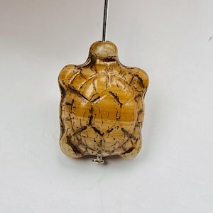 May include: A small, carved turtle bead made of brown and white agate. The bead has a detailed shell pattern and a hole drilled through the top for stringing.