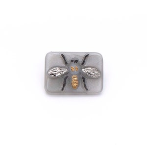 May include: A gray square button with a black and gold bee design. The bee has silver wings and a gold body.