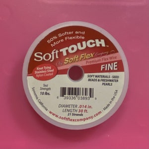 May include: A white spool of Soft Touch by Soft Flex Company Premium Flex Wire, labeled "Fine" and "Soft Materials - Seed Beads & Freshwater Pearls". The spool is 30 feet long and has a diameter of 0.014 inches. It is made in the USA and has a test strength of 10 lbs.