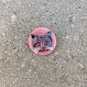 May include: A pink glass button with a black and white cat face design. The cat has a red nose and white whiskers.