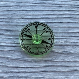 Puede incluir: Una brújula verde con una aguja negra y marcas para el norte, el sur, el este y el oeste. La brújula tiene una cubierta de plástico transparente.