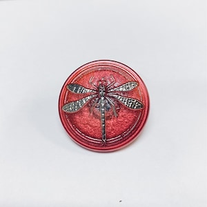 Pode incluir: Um botão vermelho com um design de libélula prateada. A libélula tem um corpo e asas detalhados.