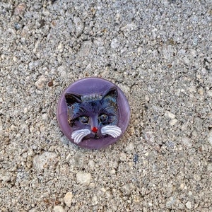 May include: A purple glass button with a black cat face, white whiskers, and a red nose.
