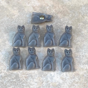 May include: A collection of grey cat-shaped buttons and a rectangular button with a metal fastener. The cat buttons are arranged in two rows of four, with the rectangular button at the top. The buttons are made of a smooth, possibly plastic material.