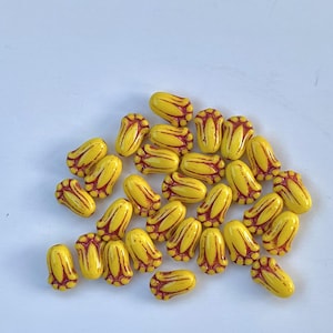 Puede incluir: Un grupo de cuentas de vidrio amarillo con detalles rojos. Las cuentas tienen forma de capullo de flor y tienen un acabado brillante.