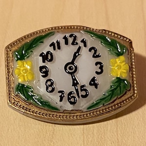 May include: A white clock face with black Roman numerals and hands, surrounded by a gold-coloured frame with green leaves and yellow flowers.