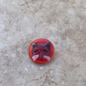 May include: A red glass button with a black cat face and white whiskers.