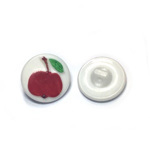May include: Two white buttons with an apple design. One button features a red apple with a green leaf and brown stem. The other button is plain white with a shank on the back. Ideal for sewing and craft projects.