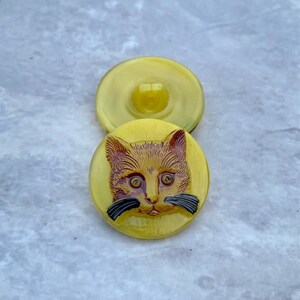 May include: A pair of yellow glass buttons with a cat face design. The cat has brown fur, black whiskers, and yellow eyes.