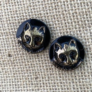 Puede incluir: Dos botones negros con un diseño de cara de gato dorado. La cara del gato está detallada con pelaje y rasgos faciales. Los botones son redondos y tienen un acabado brillante, perfectos para proyectos de costura o manualidades.