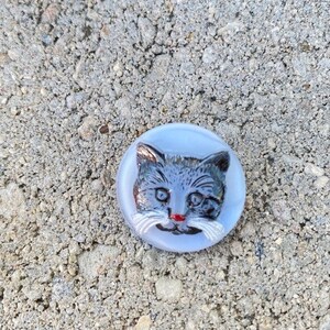May include: A white glass button with a gray cat face design. The cat has blue eyes and a red nose.