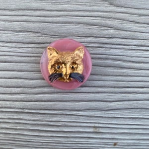 May include: A pink glass button with a gold cat face and black whiskers.