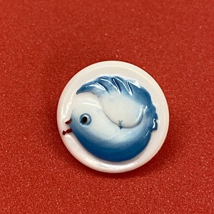May include: A white ceramic button with a blue and white bird design. The bird is facing left and has a white body with blue wings and a blue tail.