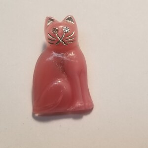 Peut inclure: Une figurine de chat en verre rose avec des détails argentés autour des yeux et des moustaches. Le chat est assis avec sa queue enroulée autour de ses pattes.
