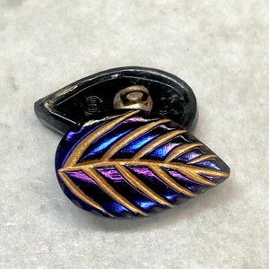 May include: A purple and gold iridescent glass button with a leaf design. The button has a black metal back with a loop for sewing.