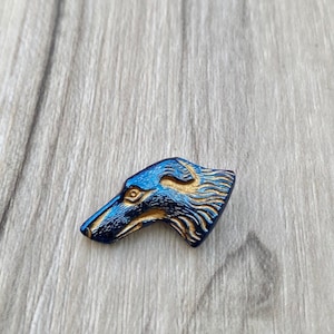 May include: A blue and gold dog head button. The button is made of a hard plastic material and has a detailed design of a dog's head. The dog's head is facing to the right and has a long snout, pointed ears, and a closed mouth.
