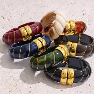 May include: A collection of chunky, colorful bracelets. The bracelets are in shades of red, blue, green, brown, black, and cream. Each bracelet features gold-colored metal accents. The bracelets are made of a glossy material.