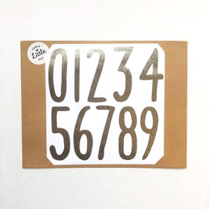 May include: A sheet of white paper with silver foil numbers 0 through 9. The numbers are in a handwritten style font.