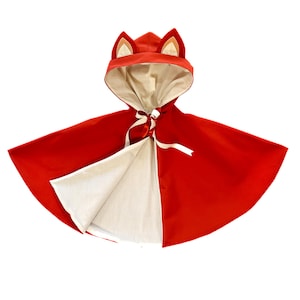 May include: A red hooded cape with fox ears. The cape is red on the outside and cream-coloured on the inside. A cream-coloured ribbon ties at the neck. The hood has fox ears with tan and cream-coloured accents. This cape is suitable for costume play.