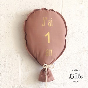 Peut inclure: Un coussin en forme de ballon de couleur rose poussiéreux avec du texte doré qui dit "J'ai 1 an". Le ballon est attaché avec une corde beige.