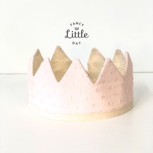 May include: A pink paper crown with a white polka dot pattern and a white satin ribbon. The crown has a pointed top and is decorated with the text "Fancy Little Day" in black.