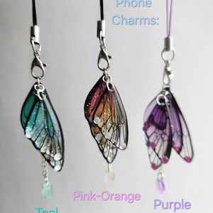 May include: Three phone charms featuring a butterfly design. The charms are teal, pink-orange, and purple. The charms are attached to black and purple phone straps. Phone Charms.