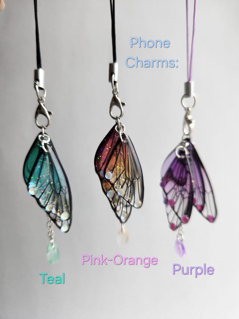 May include: Three phone charms featuring a butterfly design. The charms are teal, pink-orange, and purple. The charms are attached to black and purple phone straps. Phone Charms.