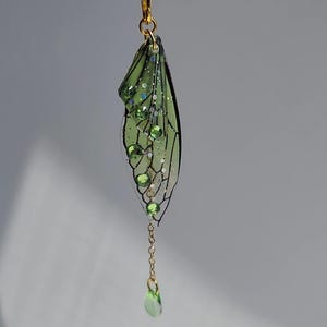 May include: A green and gold fairy wing charm with a dangling green crystal. The wing is made of clear resin with a black vein pattern and green glitter.