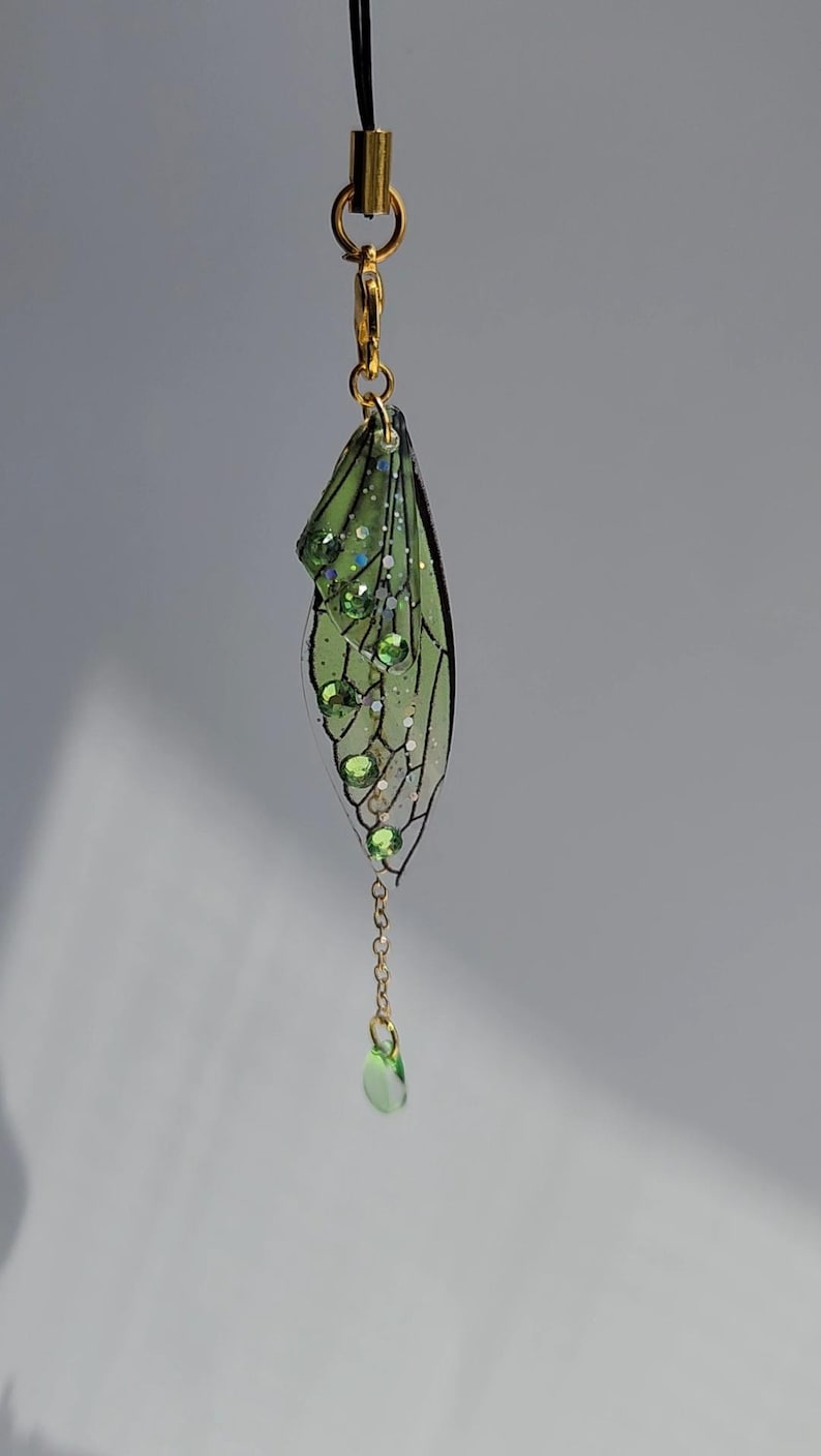 May include: A green and gold fairy wing charm with a dangling green crystal. The wing is made of clear resin with a black vein pattern and green glitter.