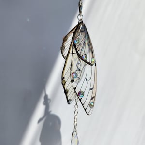 May include: A silver charm with a pair of iridescent wings and a crystal drop. The wings are translucent and have a delicate, intricate design. The charm is attached to a silver chain.