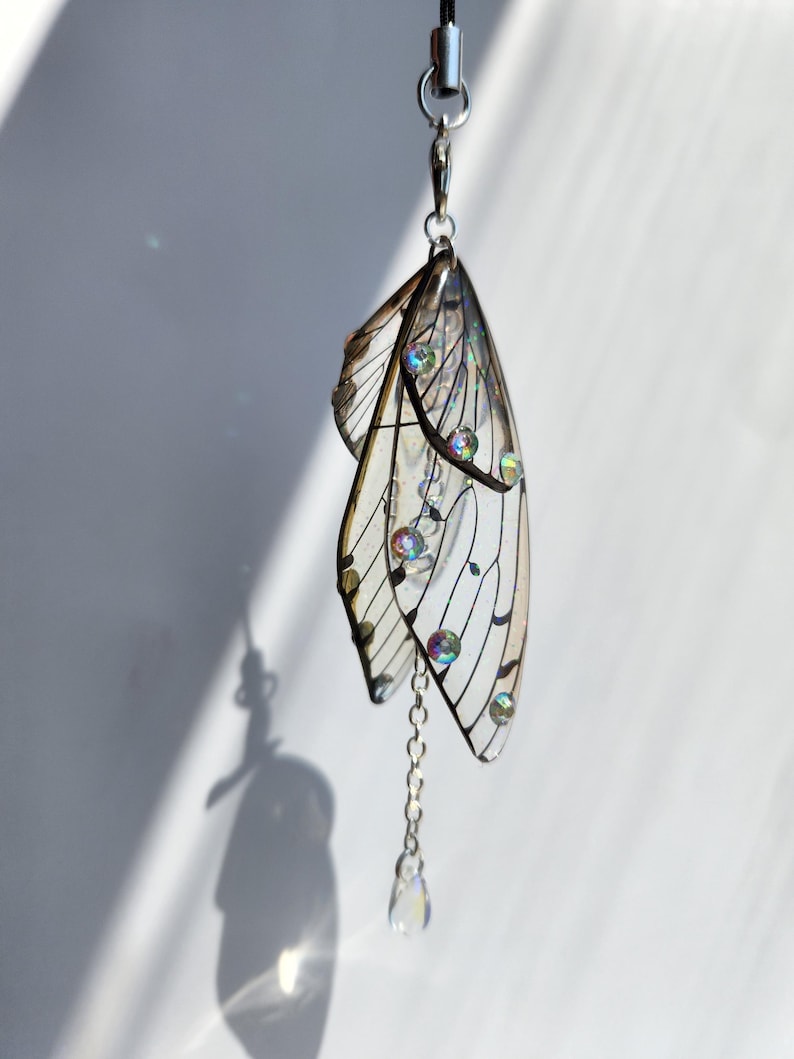 May include: A silver charm with a pair of iridescent wings and a crystal drop. The wings are translucent and have a delicate, intricate design. The charm is attached to a silver chain.