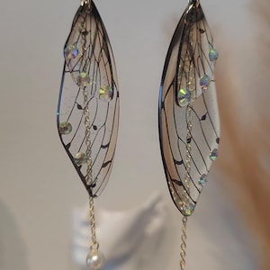 May include: A pair of dangle earrings featuring clear resin wings with black veins and iridescent rhinestones. The wings are attached to a gold chain with a pearl at the end.