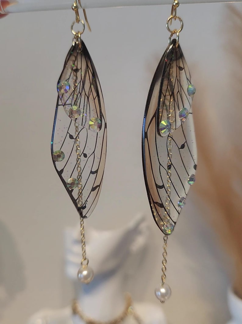 May include: A pair of dangle earrings featuring clear resin wings with black veins and iridescent rhinestones. The wings are attached to a gold chain with a pearl at the end.