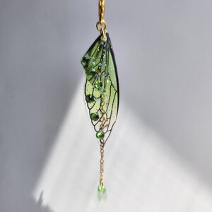 May include: A green and gold fairy wing charm with a chain and green crystal accents. The charm is hanging from a black cord.