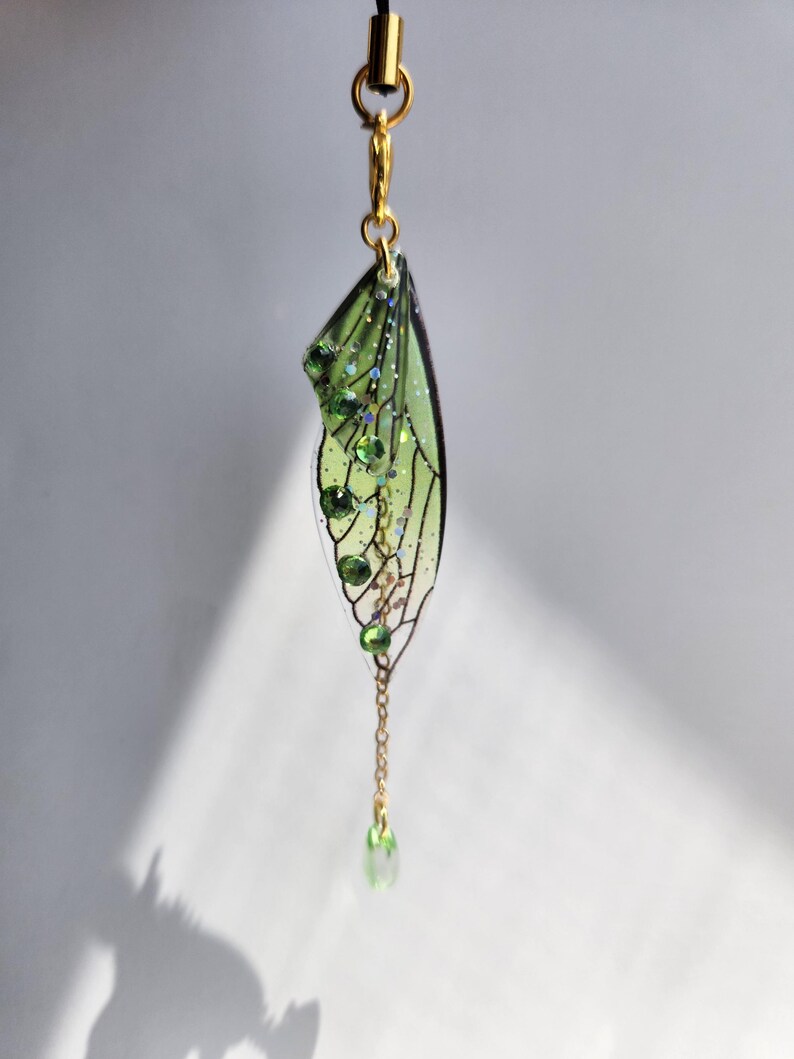 May include: A green and gold fairy wing charm with a chain and green crystal accents. The charm is hanging from a black cord.