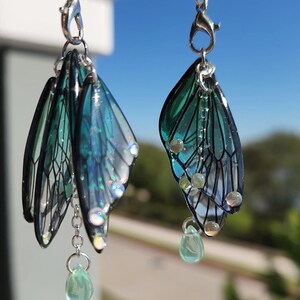 May include: A pair of dangle earrings featuring a layered design of iridescent green and blue butterfly wings with silver chain accents and green crystal drops.
