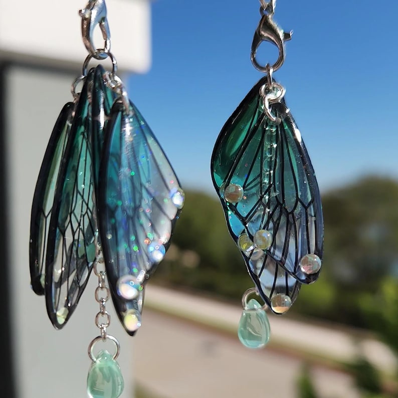 May include: A pair of dangle earrings featuring a layered design of iridescent green and blue butterfly wings with silver chain accents and green crystal drops.