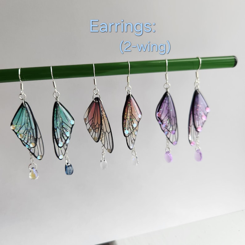 May include: Five pairs of fairy wing earrings with iridescent glitter. The wings are made of clear resin with black veins and are attached to silver hooks. The earrings are available in four colors: blue, pink, purple, and iridescent.