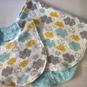 May include: A set of two baby bibs and a burp cloth with a white background and a pattern of gray, blue, and yellow clouds. The bibs are reversible with a soft blue minky fabric on one side.