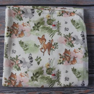 May include: A folded white blanket with a woodland animal print. The design features foxes, raccoons, and deer among floral and botanical elements in shades of brown, pink, and green. The blanket is made of soft fabric.