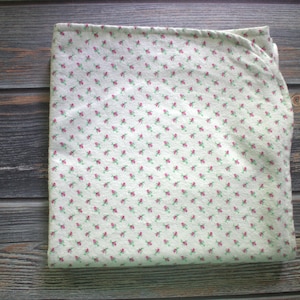 May include: Folded white fabric with a repeating pattern of small red flowers and green leaves. The fabric is set against a dark wood grain background, suggesting a textile or craft item.