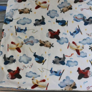 May include: White baby blanket with a repeating pattern of colorful airplanes, clouds, and stars. The airplanes are in various colors, including red, blue, and tan. The blanket has a blue trim and a blue loop.