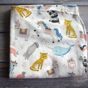 May include: White baby blanket with a colorful animal print. The blanket features illustrations of a llama, unicorn, cheetah, panda, narwhal, and other animals. The blanket is folded on a wooden surface.